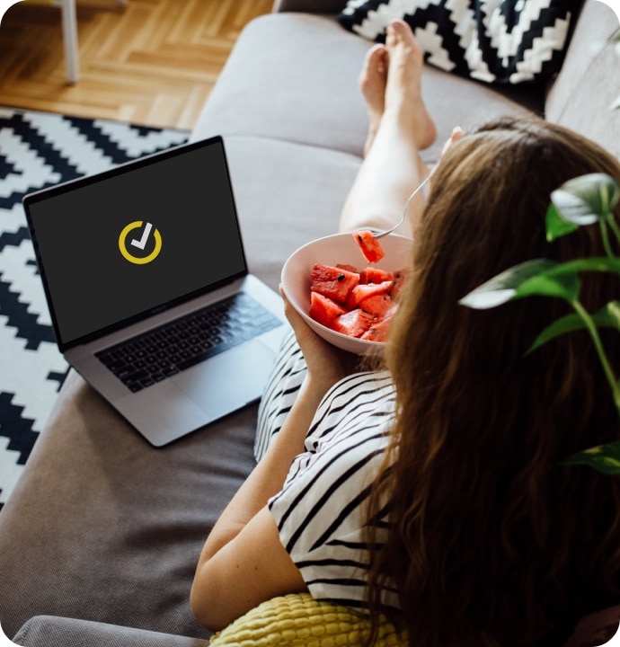 Kvinna som ligger i en soffa och äter frukt med en laptop bredvid sig med Norton-logotypen